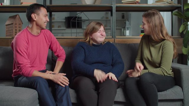 Carefree joyful diverse deaf-mute people speaking sign language, sharing funny stories and laughing sincerely on sofa at home. Cheerful hearing impaired friends enjoying leisure in domestic room.