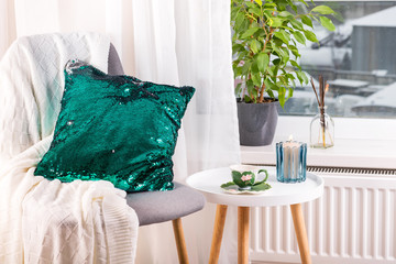 a cup of herbal tea on a table, a knitted wool blanket , green sequin pillow on a chair and candle over window . Bedroom interior