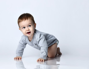 Little baby boy toddler in grey casual jumpsuit and barefoot crawling on floor, smiling and looking up over white wall background