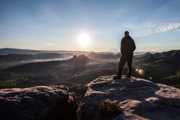 rear view of a young man standing on the top of a rock watching the sunrise above the forest - obrazy, fototapety, plakaty