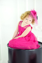 Little 4 years old girl in beautiful pink dress and pink hat with veil sitting on black barrel. Birthday girl waiting for friends and presents on birthday party. Waiting for spring small girl