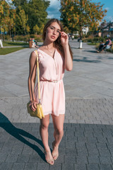 Beautiful girl in pink dress, stands in summer in city, adjusts glasses, yellow handbag, posing. Background road trees paving stones. Emotions of relaxation comfort on bright sunny day.