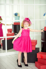 Little 4 years old girl in beautiful pink dress and pink hat with veil trying on her mother shoes near red gift boxes. Birthday girl waiting for friends on birthday party. Wait for spring.