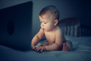 A newborn baby uses a computer while sitting on a sofa at night. The child is dressed in a diaper on his naked body.