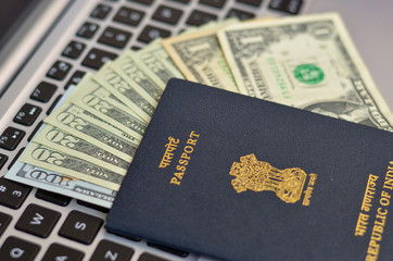 Indian Passport kept against a laptop keyboard and dollar bills, foreign currency.  National emblem, Lion Capital of Ashoka from Sarnath is embossed. Concept Wealthy IT professional Global citizen