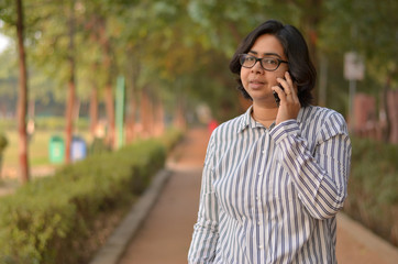 Fototapeta na wymiar Portrait of a confident looking young Indian woman with short hair and spectacles speaking on a mobile phone and holding a laptop in outdoor setting wearing a formal striped shirt in the park