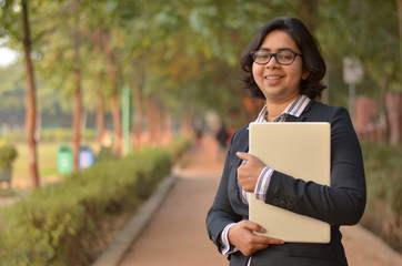 Closeup portrait of a confident young Indian Corporate professional woman with short hair and spectacles, holding a laptop in hands in an outdoor setting wearing a black white stripped shirt in a park