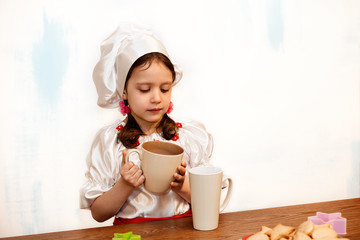 A girl in a chefs hat is standing at a kitchen table holding a cup