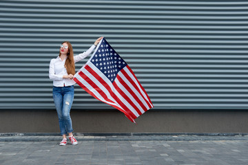 Independence day and patriotic concept. Young redhead woman with red painted lips standing with waved by wind usa flag, holding it above. Grey metal panel background.