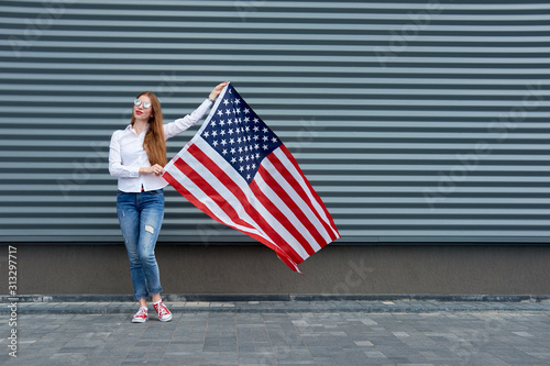Independence day and patriotic concept. Young redhead woman with red painted lips standing with waved by wind usa flag, holding it above. Grey metal panel background.