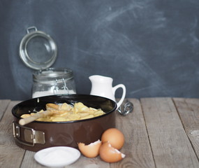 On the kitchen cutting board baking dish with dough. Ingredients and kitchen utensils nearby on an ancient wooden table.