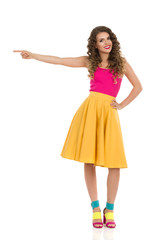 Cute Young Woman In Colorful High Heels, Vibrant Skirt And Pink Top Is Pointing At The Side.