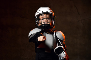 Boy in hockey uniform and protective helmet with hockey skates standing and pointing on camera with...