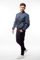 Young european man in white sweater and black pants, blue bomber jacket posing on white background....