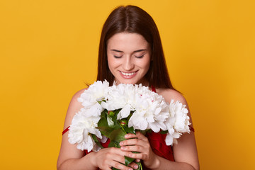 Obraz na płótnie Canvas Indoor shot of young cute woman smelling white flowers,looking down at her bouquet, holding gentle peonies in hands, attractive smiling lady posing with flowers against yellow studio background.