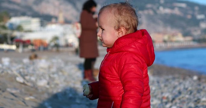 Cute Blond Baby Boy (Two Years Old) In A Red Down Jacket Eating A Croissant On The Beach, French Riviera. Close Up Portrait - DCi 4K