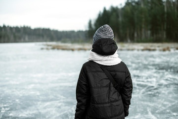 Fototapeta na wymiar Young woman in snow blizzard standing alone on the lake ice and staring forward. Girl's back. Cold atmosphere in the afternoon. Foggy air. Winter concept.