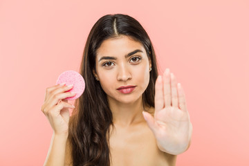 Young indian woman holding a facial skin care disc standing with outstretched hand showing stop...