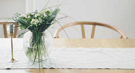 Close view of fresh flowers in glass vase standing on a wooden table in white dining room interior. Interior design in minimal style.