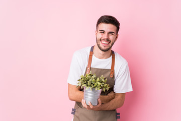 Young caucasian gardener man holding a plant isolated laughing and having fun.