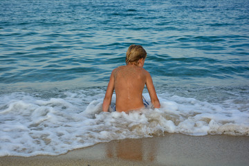Fototapeta premium A blond boy from behind, sitting in a wave on a sandy beach