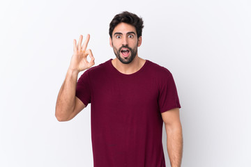 Fototapeta premium Young handsome man with beard over isolated white background surprised and showing ok sign