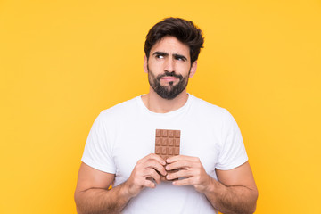 Young handsome man with beard over isolated yellow background taking a chocolate tablet and having...