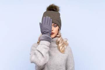 Young teenager girl with winter hat over isolated blue background covering a eye by hand