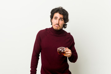 Young man holding a tea cup shrugs shoulders and open eyes confused.