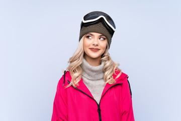 Skier teenager girl with snowboarding glasses over isolated blue background laughing and looking up
