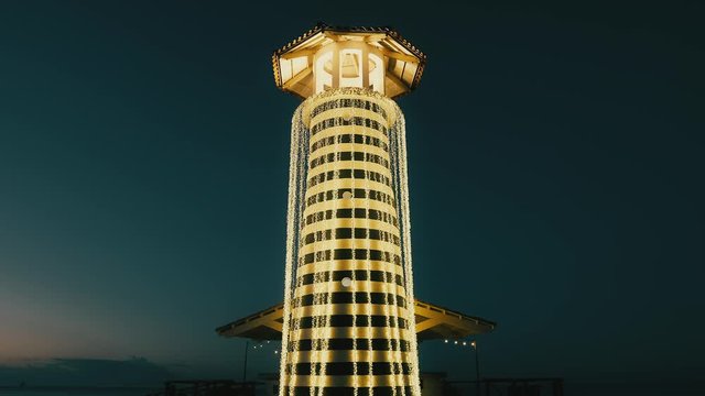 Beautiful sea lighthouse against the blue night sky. Navigation tower on summer tropical Caribbean beach. Striped pole with luminous garlands.