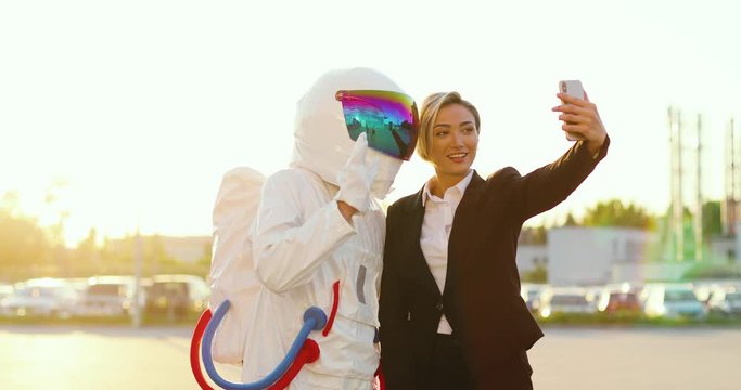 Caucasian beautiful woman in business outfit and astronaut man taking selfie photo together on the smartphone camera while being outdoors at the parking on a sunny day.
