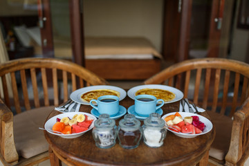 A wooden table is set on the terrace of the room. Balinese tropical breakfast of fruit, coffee and scrambled eggs and banana pancake for two. on the street near the pool