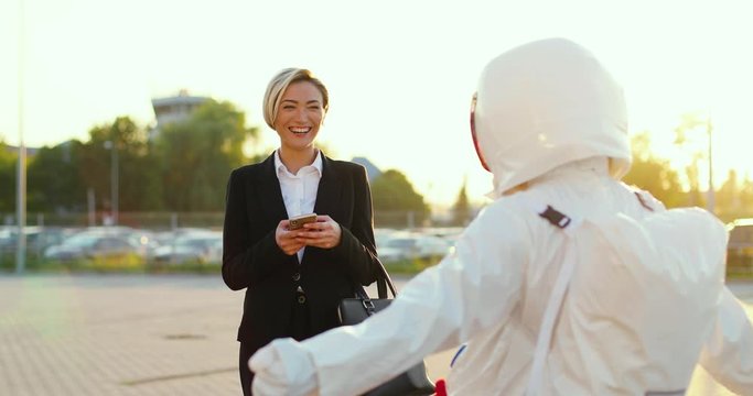 Caucasian attractive young businesswoman tapping and texting on the smartphone while standing at the parking outdoors and man in the costume of astronaut making funny things so she laughing.