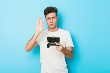 Young caucasian man playing videogames with phone standing with outstretched hand showing stop sign, preventing you.