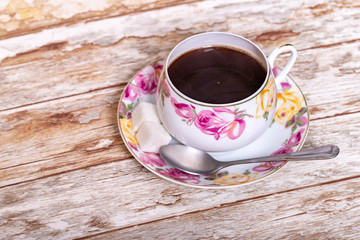 White porcelain Cup with a delicate pattern of pink and yellow rose buds with instant coffee with a spoon and two pieces of sugar on a saucer on a wooden background.