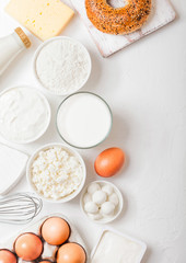 Fresh dairy products on white table background. Glass of milk, bowl of flour and cottage cheese and eggs. Fresh baked bagel with knife. Steel whisk. Top view