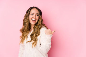 Young curvy woman posing in a pink background isolated points with thumb finger away, laughing and...