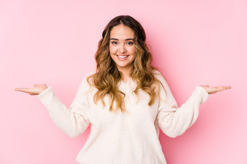 Young curvy woman posing in a pink background isolated makes scale with arms, feels happy and confident.