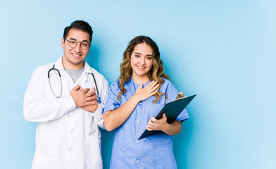 Young doctor couple posing in a blue background isolated has friendly expression, pressing palm to...
