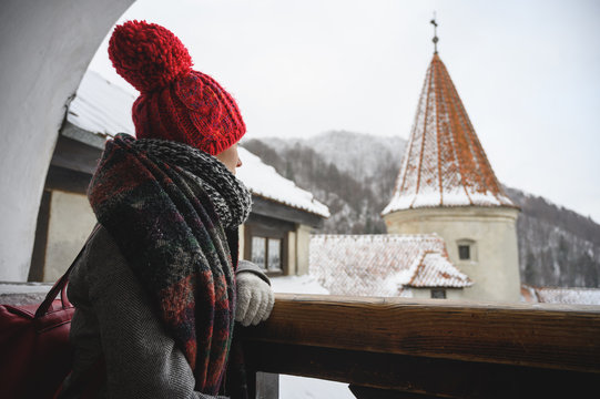Beautiful young woman watching a medieval castle, sitting on a valcon, dressed in winter clothes, scarf, hat and coat watching the landscape