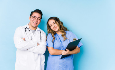 Young doctor couple posing in a blue background isolated laughing and having fun.