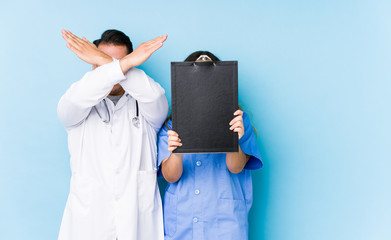Young doctor couple posing in a blue background isolated keeping two arms crossed, denial concept.