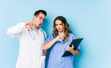 Young doctor couple posing in a blue background isolated showing thumb down and expressing dislike.