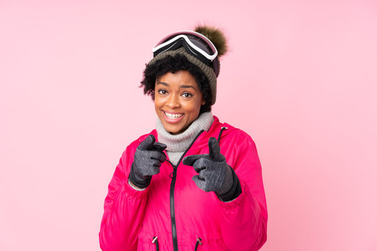 African american skier woman with snowboarding glasses over isolated pink background points finger at you