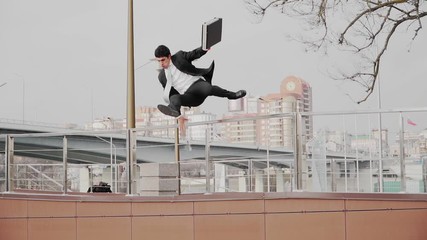 A guy in a business suit and tie with briefcase in hand, jumps over the parapet. A businessman rushing to a meeting and uses the parkour. Slow motion - Powered by Adobe