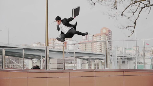 A guy in a business suit and tie with briefcase in hand, jumps over the parapet. A businessman rushing to a meeting and uses the parkour. Slow motion