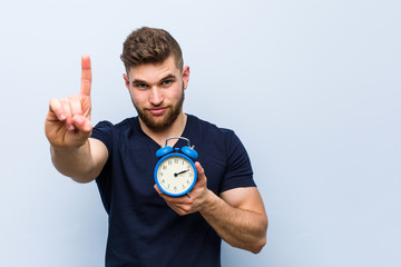Young caucasian man holding alarm clock showing number one with finger.