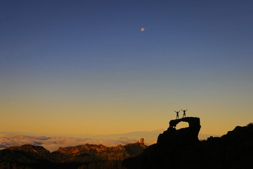 Fototapeta na wymiar Two silhouettes victoriously standing on a rock arch on a mountain top with clouds below during yellow sunrise. Roque Nublo and Teide mountain in the background. Moon on the blue sky. Canary Islands