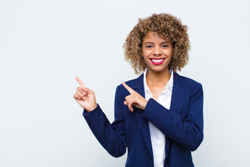 young woman african american smiling happily and pointing to side and upwards with both hands showing object in copy space against flat wall - obrazy, fototapety, plakaty